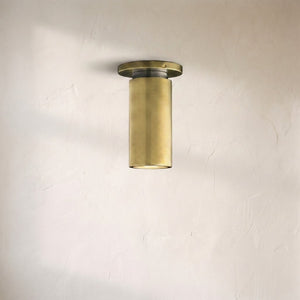 A modern cylindrical wall sconce with a brushed brass finish, mounted on a textured off-white plaster wall.