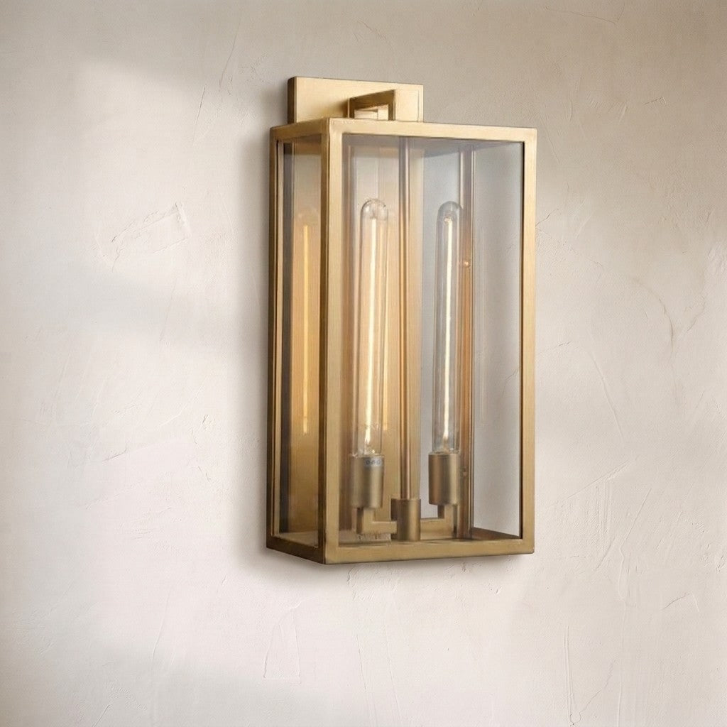 A modern wall-mounted light fixture with a brushed brass frame and clear glass panels, featuring two vertical exposed filament bulbs, mounted on a textured off-white wall.