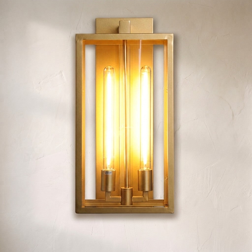 A modern rectangular wall sconce with a brushed gold metal frame and two glowing vertical filament bulbs, mounted on a textured off-white wall.