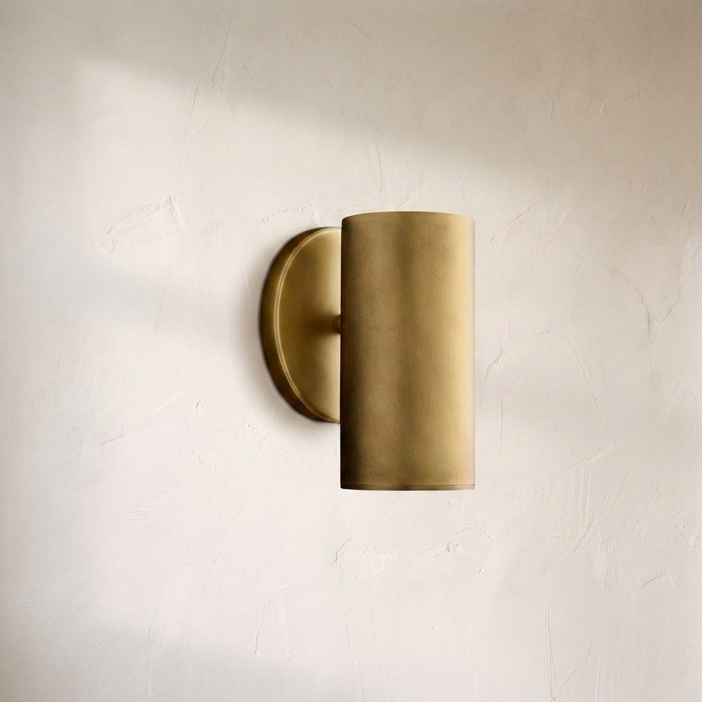 A modern cylindrical wall sconce in a brushed brass or gold finish, mounted on a textured off-white plaster wall.