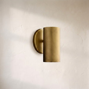 A modern cylindrical wall sconce in a brushed brass or gold finish, mounted on a textured off-white plaster wall.