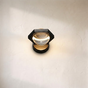 Eldric wall sconce on a plaster wall, black hexagonal frame around a clear glass lens with a warm LED halo